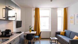 Une cuisine moderne dotée d’un four encastré, un coin repas avec une table et des chaises, un canapé avec des coussins et une fenêtre ornée de rideaux jaunes.