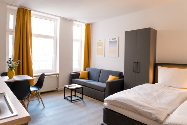 A modern hotel room with a bed, a sofa, a small table, and a desk. There are two windows with curtains, and the room has a neutral color scheme.