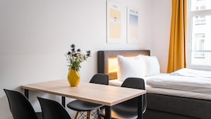 Hotellihuone, jossa on sänky, yöpöytä lampun kanssa, pieni pöytä tuoleineen sekä maljakko kukkineen.