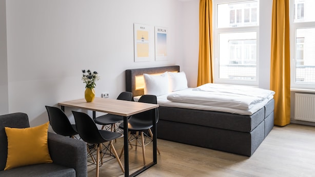 Moderni hotellihuone, jossa on sänky, työpöytä tuoleineen, maljakko kukkineen sekä kaksi kehystettyä kuvaa seinällä.