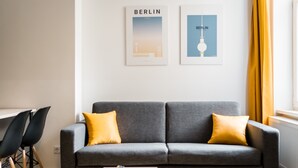 A modern living room with a grey sofa, two yellow cushions, a remote control on a small table, and two framed pictures on the wall. - Boxi Studios Berlin S (Berlin)