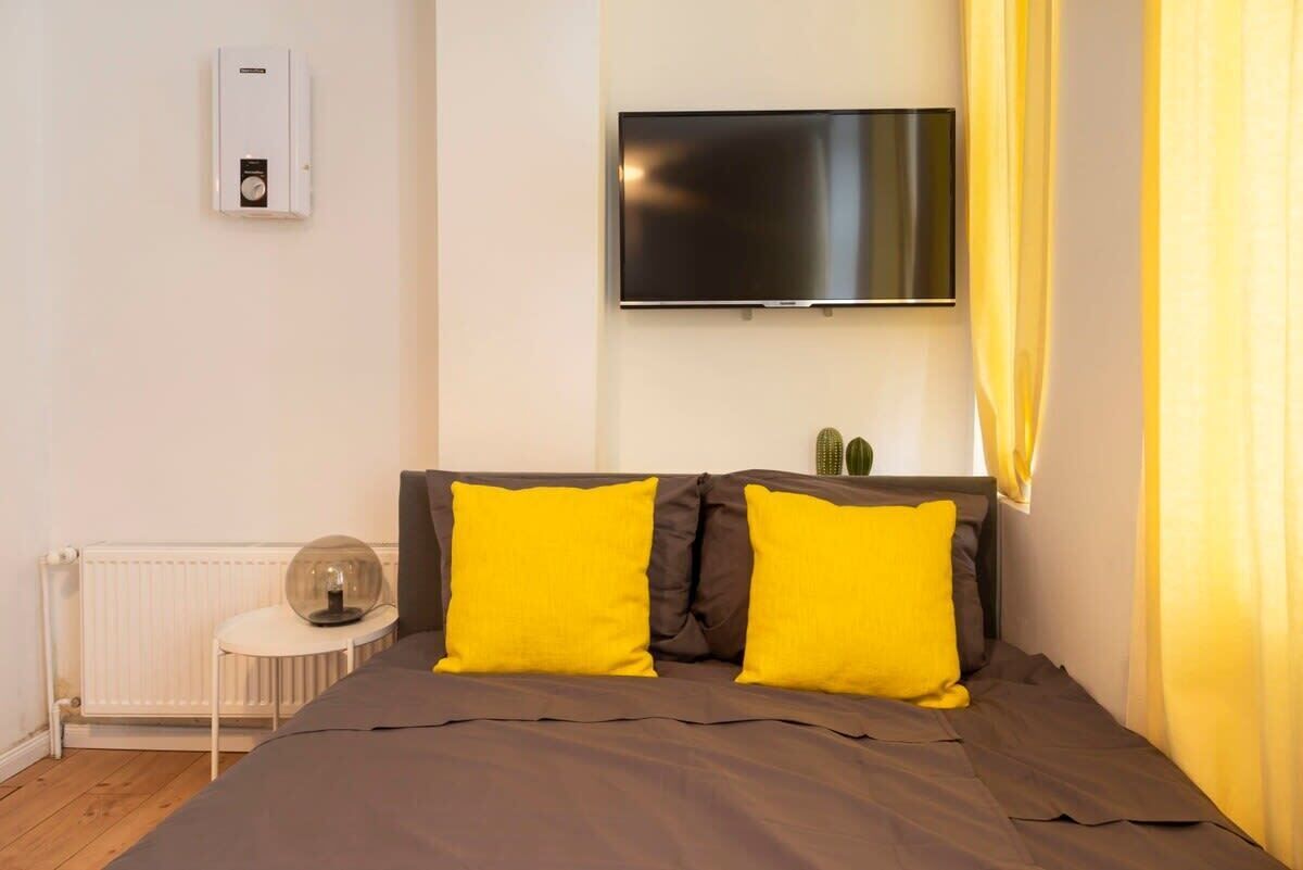 A bedroom with a bed, a TV mounted on the wall, a radiator, and a small table with a glass dome.