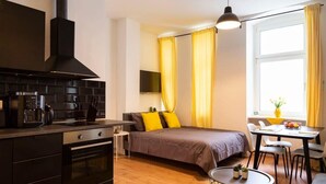 A compact room with a kitchenette, a bed with yellow pillows, a dining area with a table and chairs, and a window with curtains. - Lovely Apartment in the near of Kudamm (Berlin)
