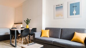 Ein modernes Wohnzimmer mit grauem Sofa, gelben Kissen, einem Holztisch und einem Essbereich mit Tisch und Stühlen. An der Wand hängen zwei gerahmte Bilder von Berlin.