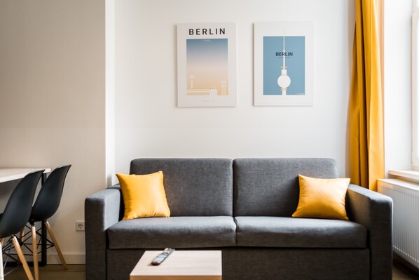 Un salon moderne avec un canapé gris, deux coussins jaunes, une télécommande posée sur une petite table et deux tableaux encadrés accrochés au mur.