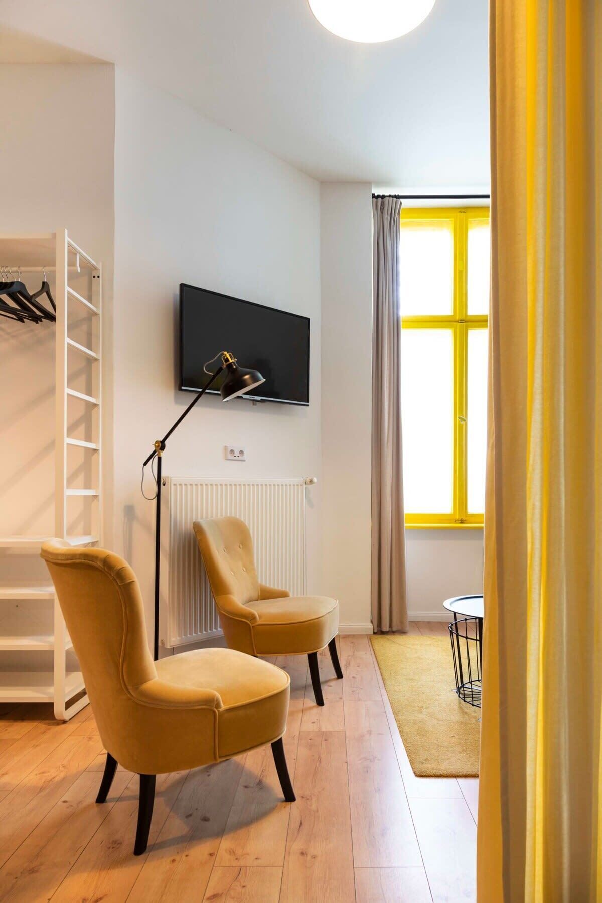 A modern living room with a flat-screen TV mounted on the wall, a floor lamp, two armchairs, a radiator, and a window with curtains.