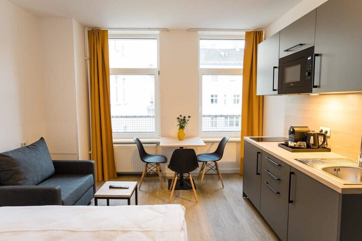 A compact hotel room with a bed, a small table, two chairs, a kitchenette, and a window with curtains.