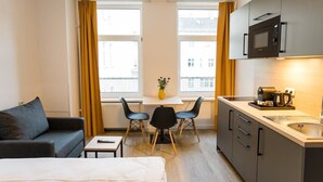 A compact hotel room with a bed, a small table, two chairs, a kitchenette, and a window with curtains.