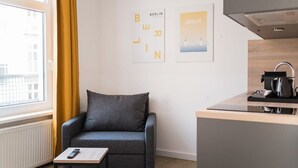 A modern room with a grey sofa, a small table, a window with yellow curtains, a kitchen area, and two framed pictures on the wall.