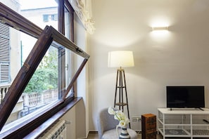 Une pièce avec une grande fenêtre, un lampadaire, un téléviseur posé sur un meuble blanc et une chaise.