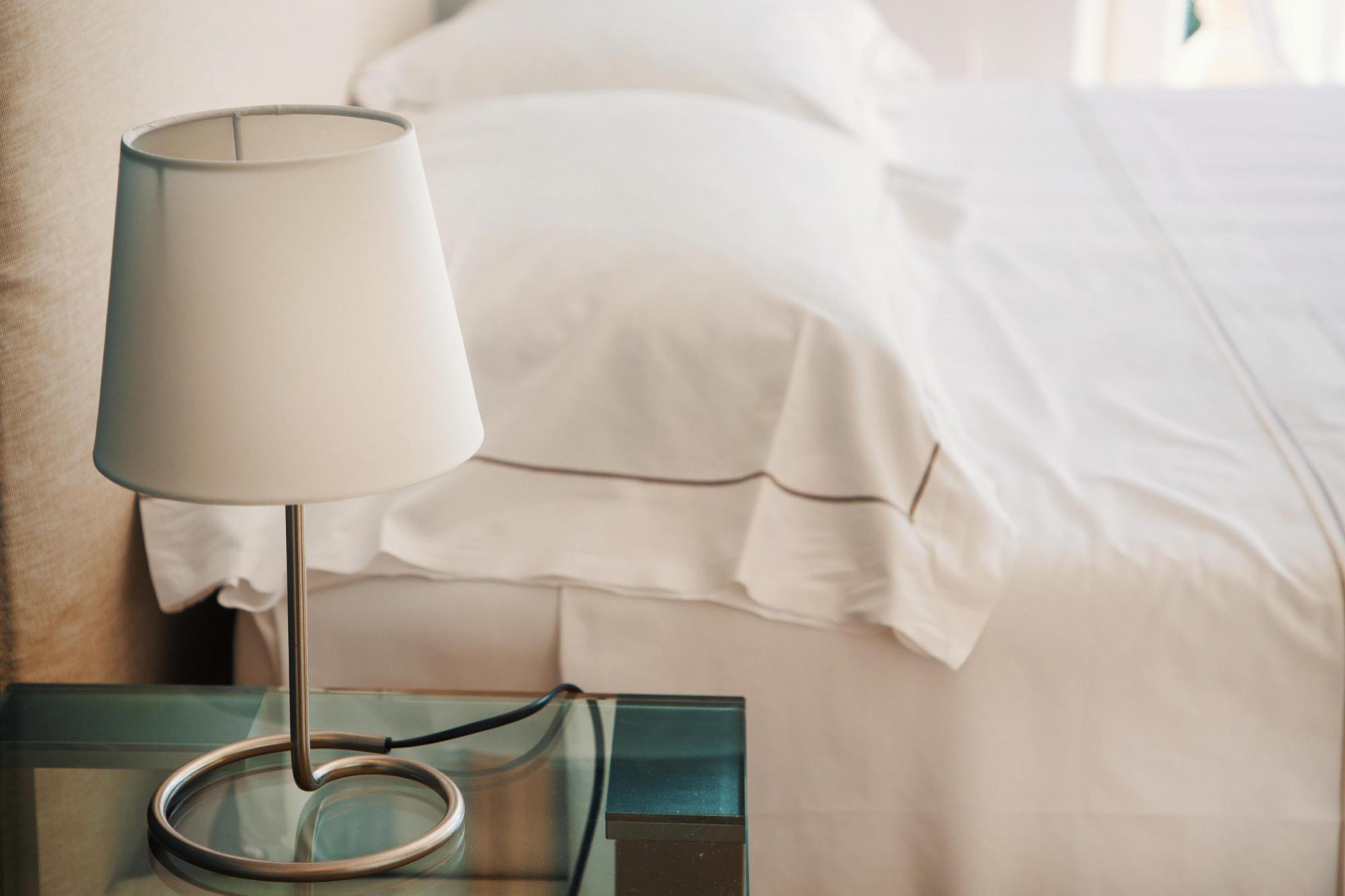 Un lit avec du linge de lit blanc, une table de chevet en verre avec une lampe, et un mur de couleur beige.