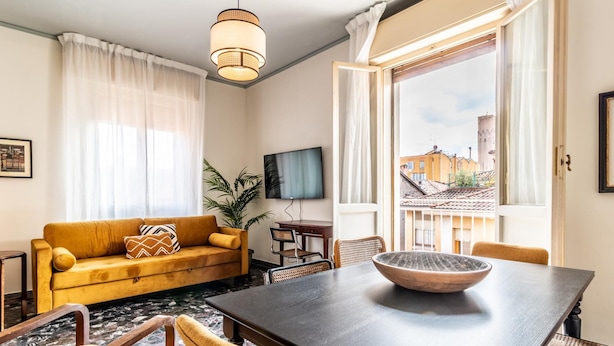 A cozy living room with a sofa, a dining table with a bowl, a TV, and a view of buildings through a window. - Dimora Vintage San Tommaso by Wonderful Italy (Bologna)