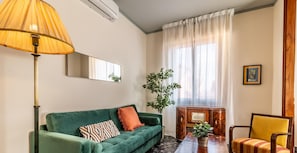 Un salon avec un canapé vert, un fauteuil jaune, une table d’appoint en bois, une lampe et une grande fenêtre ornée de rideaux légers.