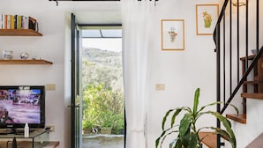 A living room with a TV, shelves with books and decorative items, a staircase, and a view of greenery outside. - Villa Rica by Wonderful Italy (Santa Margherita Ligure)