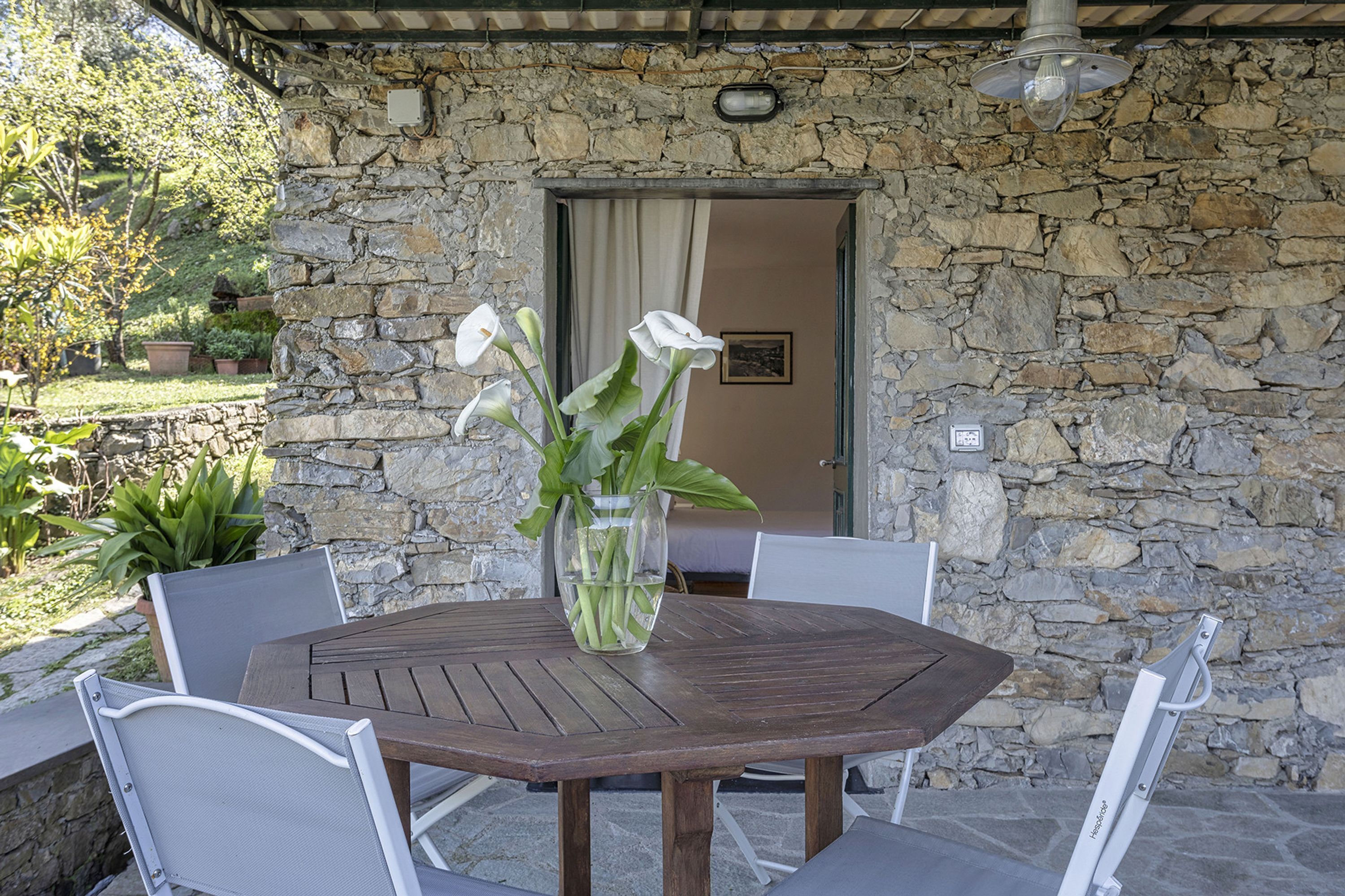Pared de piedra exterior con una mesa y sillas de madera, un jarrón con flores y una puerta que da al interior.