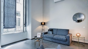 A modern living room with a grey sofa, a round mirror, a floor lamp, and a small table with a picture frame.