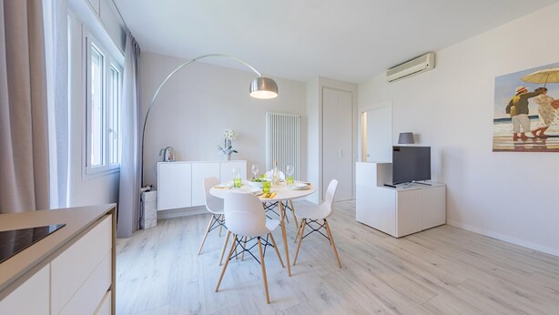 Eine moderne Küche mit weißen Schränken, einem Esstisch mit Stühlen, einer Pendelleuchte und einem Fernseher auf einem Schrank.
