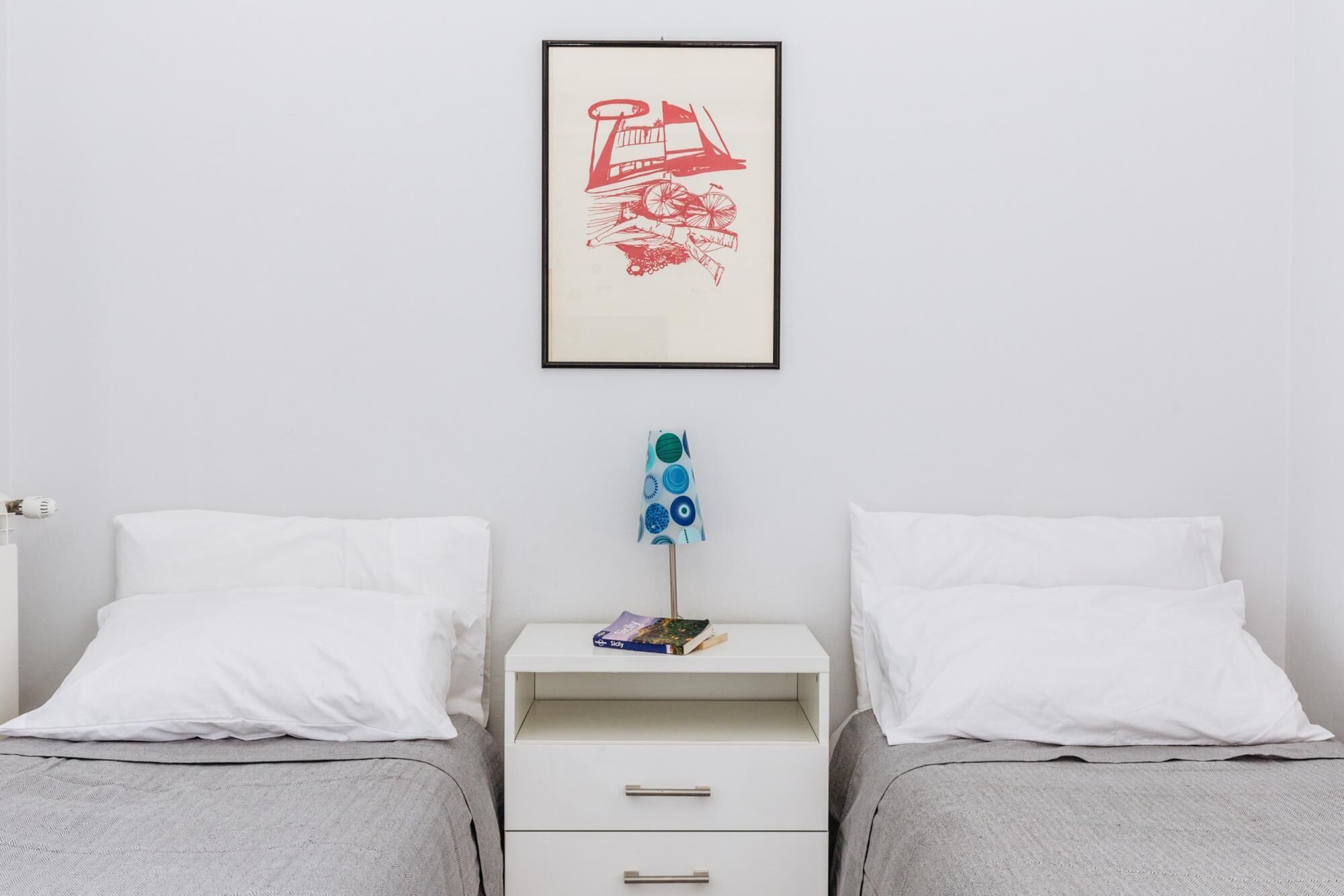 Een slaapkamer met twee enkele bedden, een witte nachtkastje met lades, een lijst met een afbeelding aan de muur en een lamp met een blauw geborduurd lampscherm.