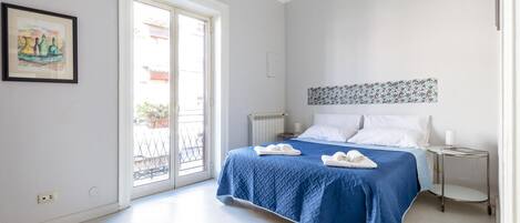 A bedroom with a bed, a bedside table, a radiator, a window, and a door.