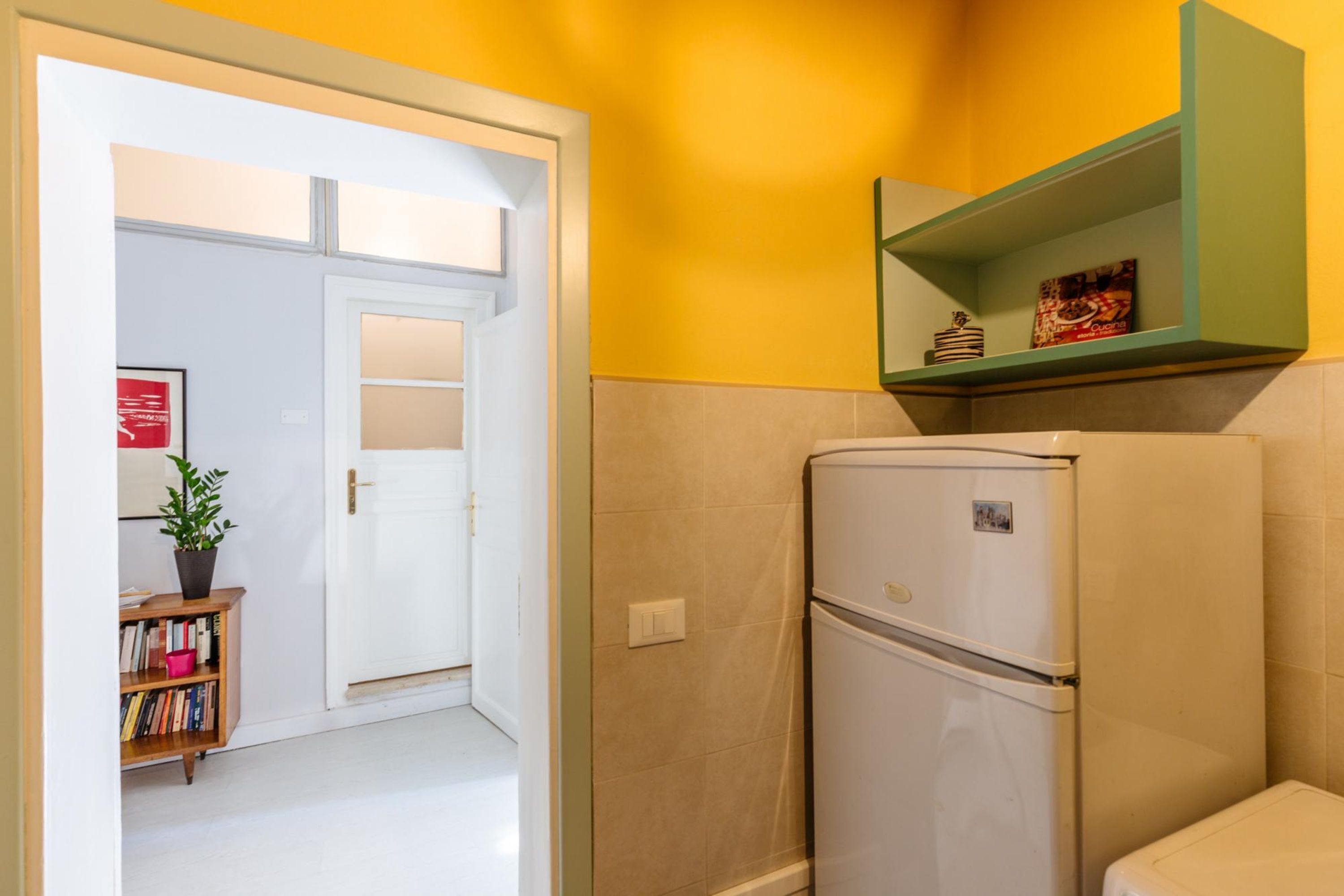 A kitchen with a white refrigerator, a green shelf above it, and a door leading to another room.