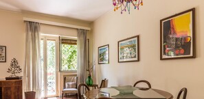 Un comedor con una mesa redonda, sillas y una araña. Hay cuadros en la pared y una ventana con cortinas.