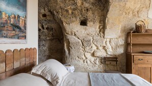 A bedroom with a stone wall, a wooden headboard, a bed with white linens, and a painting of a cityscape. - Casa Petra by Wonderful Italy (Modica)