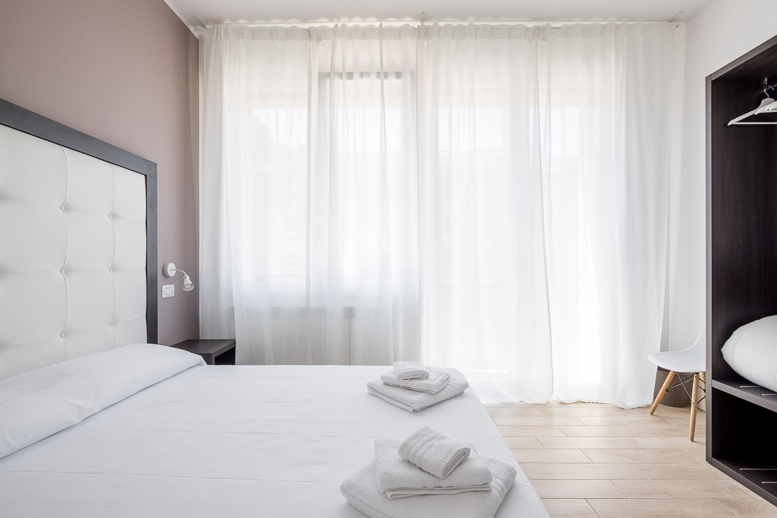 A modern hotel room with a large bed, a bedside table, a wardrobe, and a window with sheer curtains.