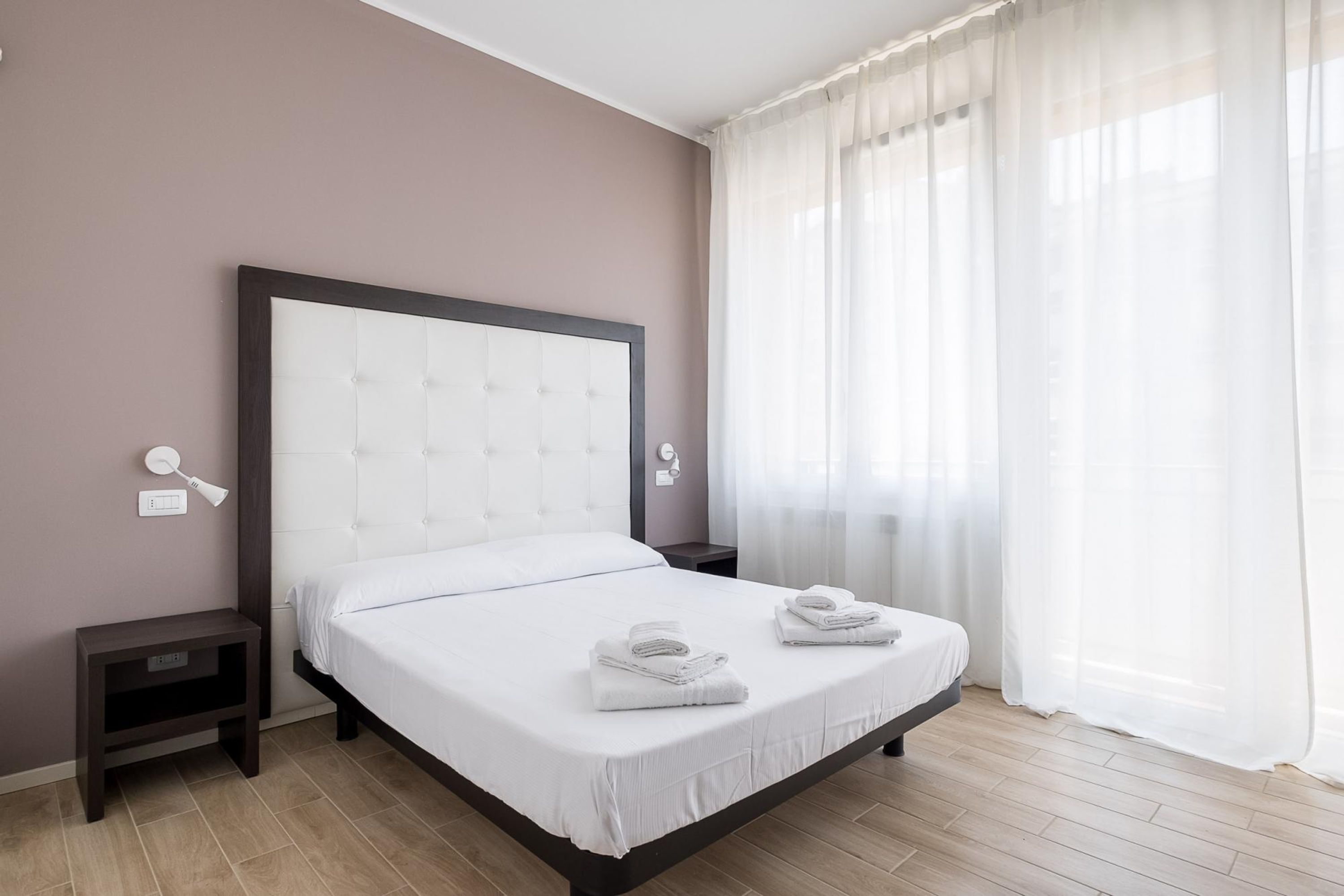 A neatly made bed with white linens and two folded towels, a bedside table with a lamp, and a window with sheer curtains.
