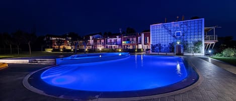 Piscina iluminada em azul, com borda e um terraço de tijolos ao redor. Ao fundo, um edifício moderno com design de fachada único e varanda.