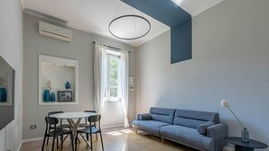 Een moderne woonkamer met een grijze bank, een ronde eettafel met stoelen, een grote spiegel en een plafondlamp.