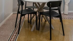 Een eetruimte met een witte tafel, vier zwarte stoelen, een witte airconditioner, een raam met een witte omlijsting en uitzicht op een gebouw.