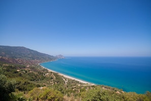 A coastal landscape with a beach, clear blue sea, and surrounding greenery.