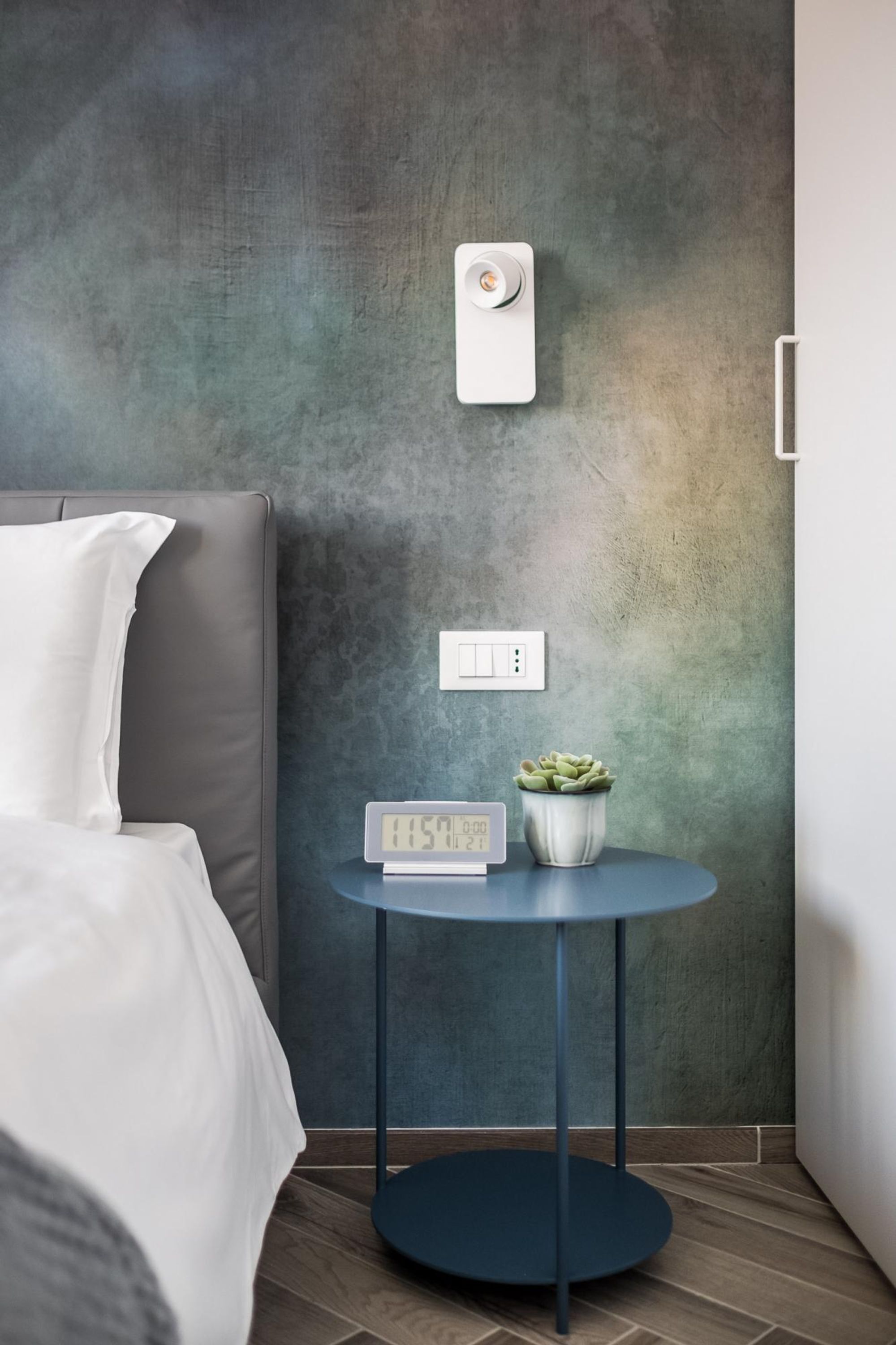 Quarto moderno com cama cinza, mesa lateral redonda azul com relógio digital, interruptor de luz na parede e um pequeno vaso de planta.