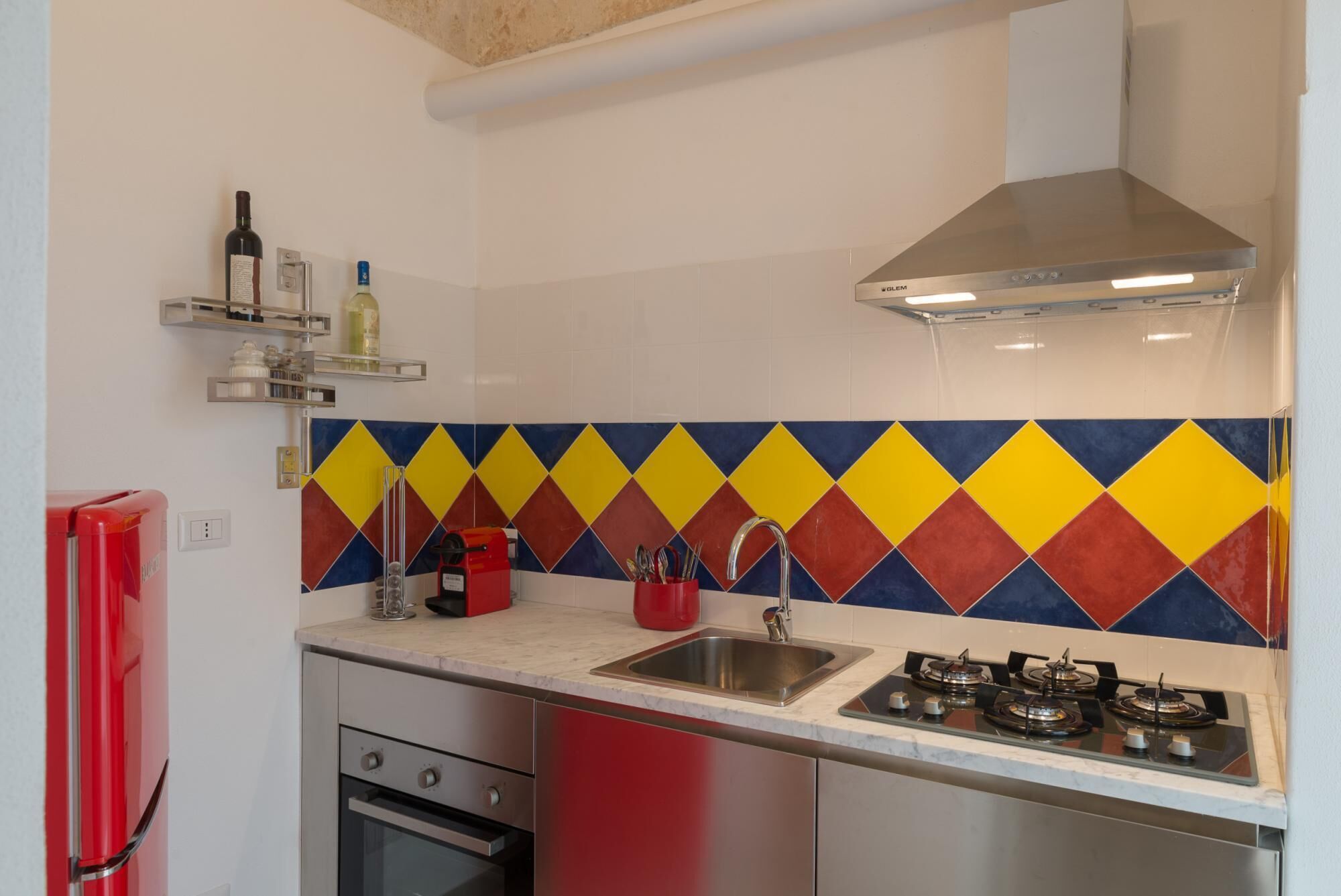 Eine Küche mit farbigem, diamantförmigem Fliesenspiegel, einem Edelstahl-Spülbecken, einem Gasherd und einem roten Kühlschrank.
