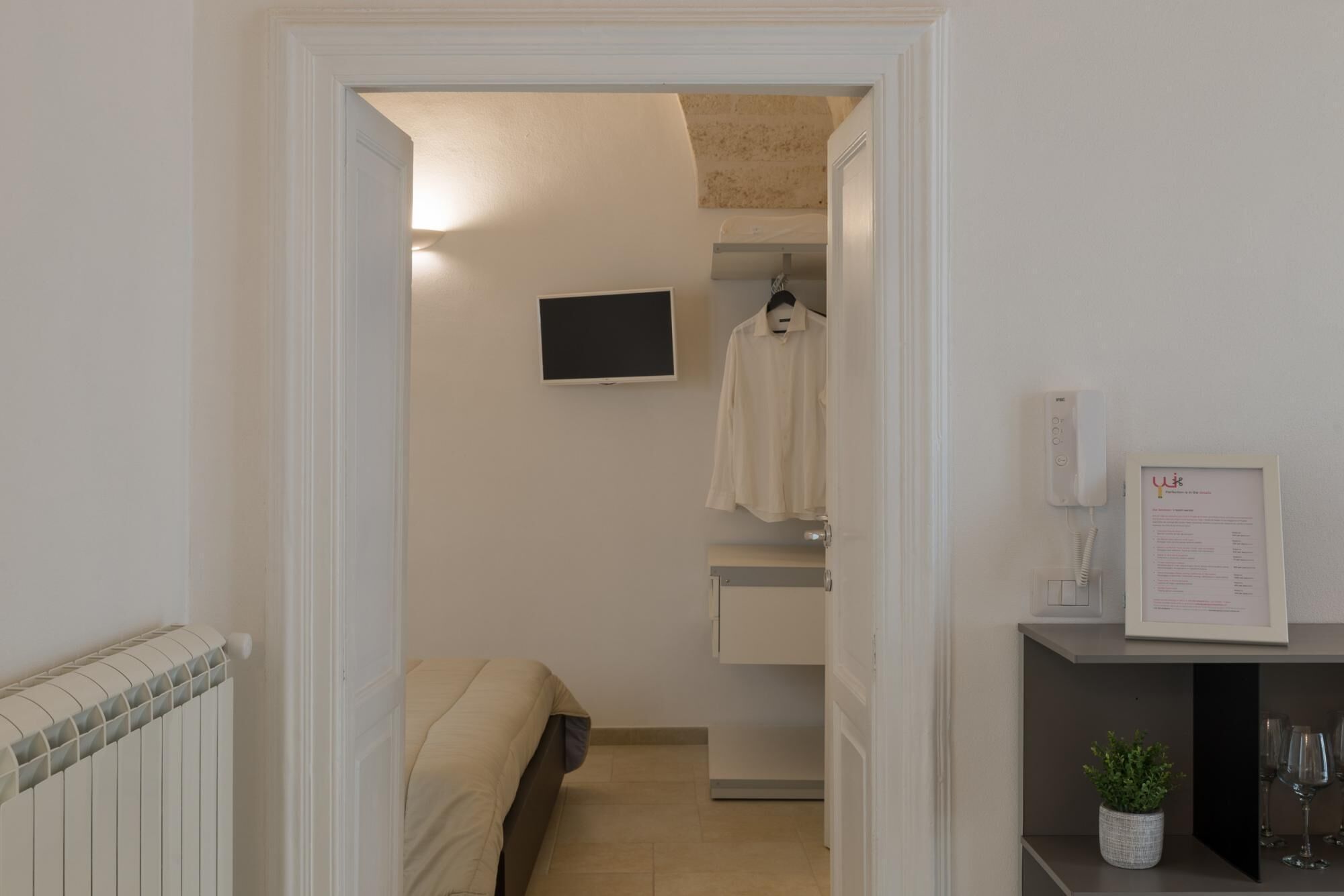 A hotel room with a bed, a TV mounted on the wall, a coat hanging, a bedside table with a plant, and a framed menu on the wall.