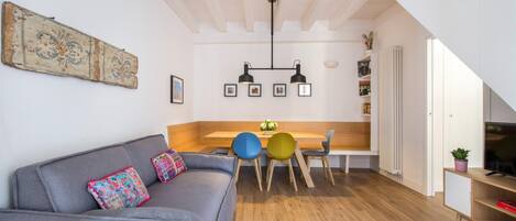 Una sala de estar moderna con un sofá gris, cojines de colores, una mesa de comedor de madera con sillas y una zona de cocina con una lámpara colgante.
