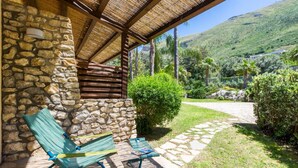 Un bâtiment en pierre surmonté d’un toit de chaume, deux fauteuils de jardin verts et un chemin pavé menant à un paysage verdoyant luxuriant, avec des palmiers et une montagne en arrière-plan.