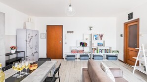 Ett modernt vardagsrum med trägolv, en grå soffa, ett matbord med vinglas och en bokhylla med böcker och dekorativa föremål.