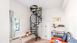 A bedroom with a spiral staircase, a bed with patterned bedding, a small shelf with a radio, and a window with curtains. - Gaudio Flat Near Ariston Theater by Wonderful Italy (Sanremo)