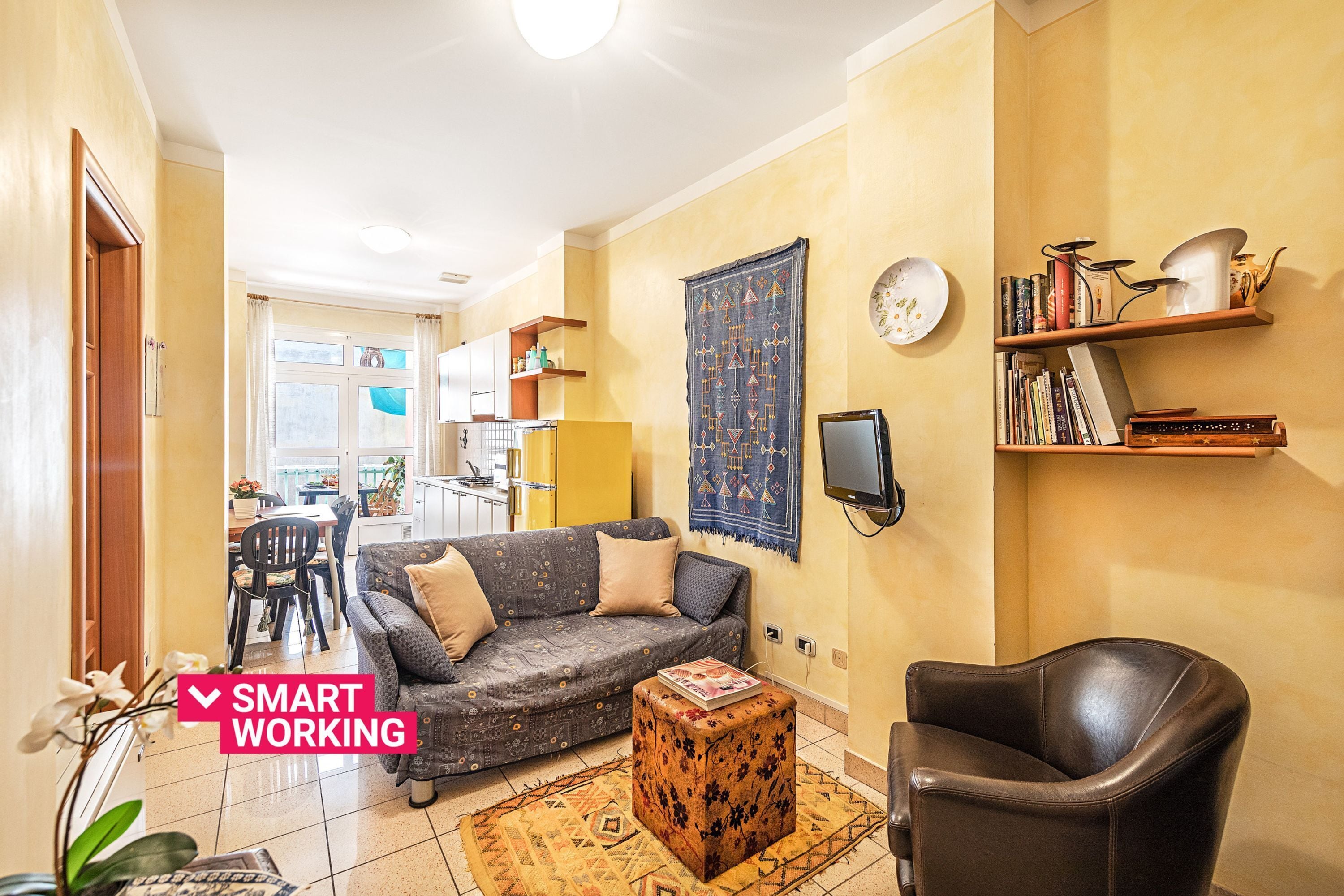 Un salon comprenant un canapé, un pouf et un fauteuil, un téléviseur fixé au mur, ainsi qu’une bibliothèque remplie de livres et d’objets décoratifs.