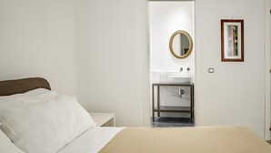 A bedroom with a bed, a bedside table, a bathroom with a sink and mirror, and a framed picture on the wall. - Mirabella Apartment in Ortigia by Wonderful Italy (Syracuse)