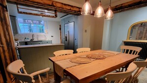 Un coin repas avec une table et des chaises en bois, une cuisine en arrière-plan et des luminaires suspendus au plafond.