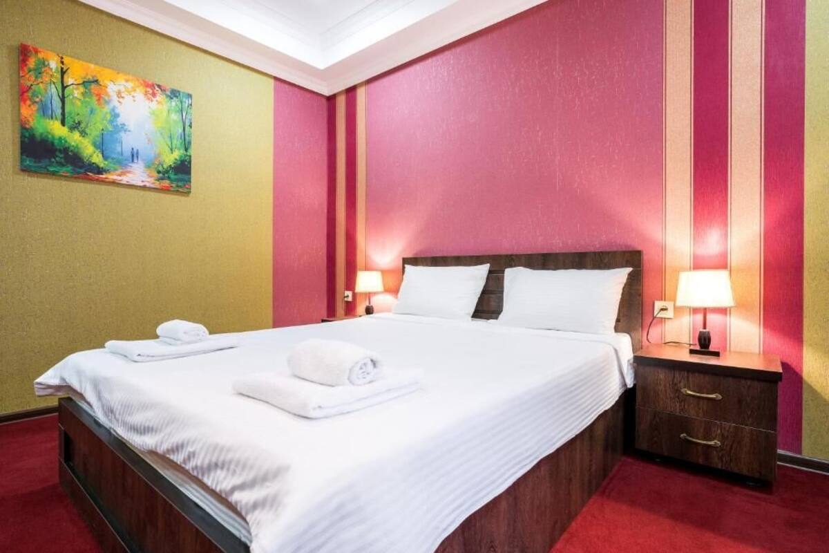 A hotel room with a large bed, two bedside tables with lamps, a painting on the wall, and a red carpet.