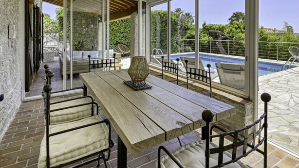 Une terrasse ensoleillée avec une table et des chaises en bois, une piscine et une vue sur la verdure.