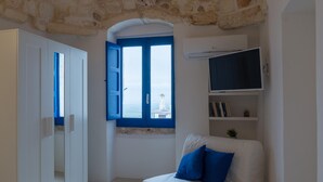 Quarto com sofá branco, janela azul, guarda-roupa branco e televisão na parede.
