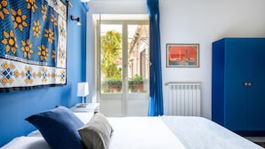 A hotel room with a bed, a nightstand, a blue wardrobe, a window with a view of buildings, and a patterned curtain. - Iconic Apt in Porta Carini by Wonderful Italy (Palermo)