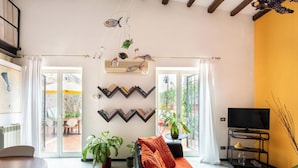 A modern living room with a sofa, a coffee table, and a TV stand. There are plants and a wall-mounted shelf with books. - Iconic Apt in Porta Carini by Wonderful Italy (Palermo)