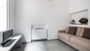A minimalist living room with a flat-screen TV on a stand, a white air conditioning unit, a beige sofa with two cushions, and a shelf with decorative items. - San Michele Apartments - Lignum by Wonderful Italy (Bologna)