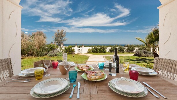 A dining table set for two with plates, cutlery, and glasses, overlooking a garden and the sea. - Villetta Tisbe by Wonderful Italy (Ostuni)