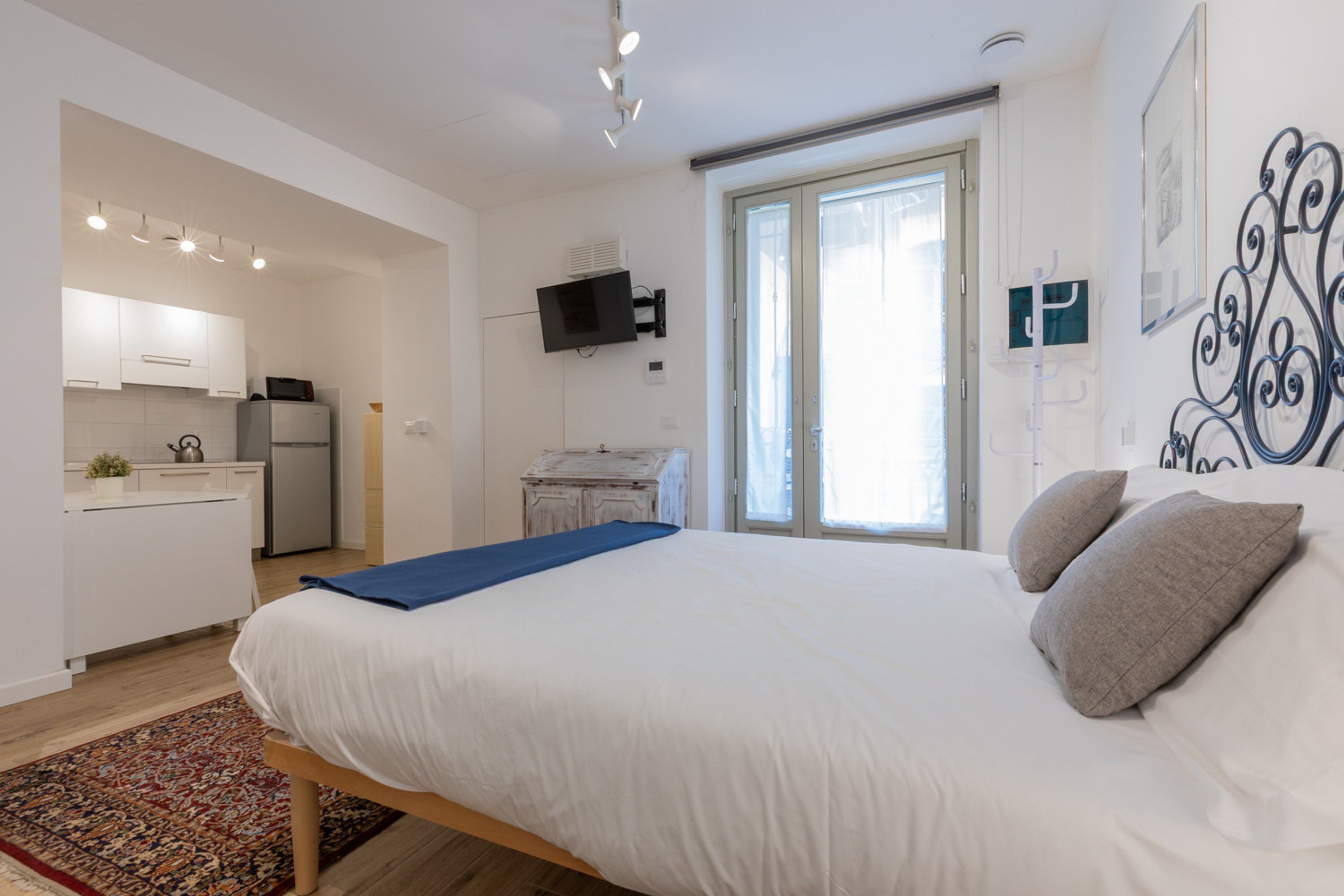 A bedroom with a large bed, a small kitchen area, and a sliding glass door leading to a balcony.